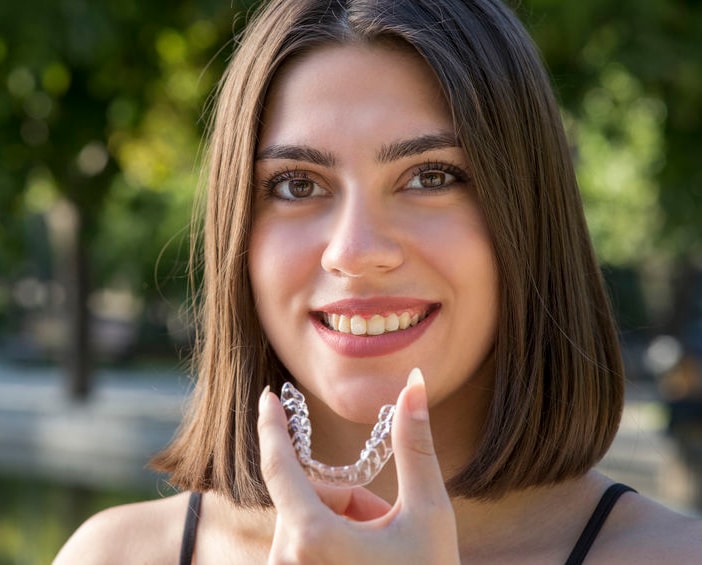 A girl smiling and holding up a clear aligner in front of some trees.