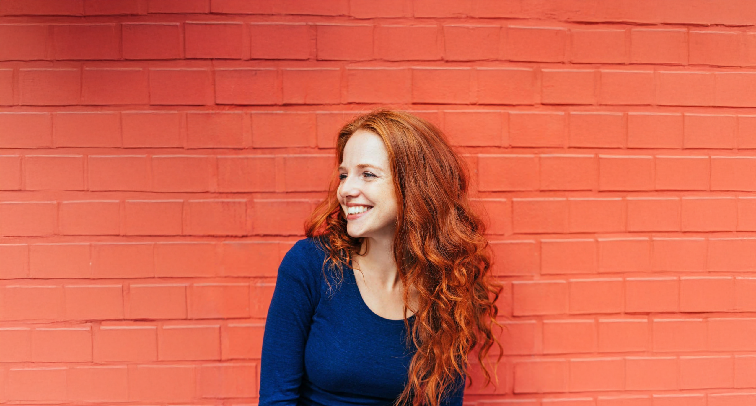 A woman in a dark blue shirt smiles against a brick wall background, looking to her right.