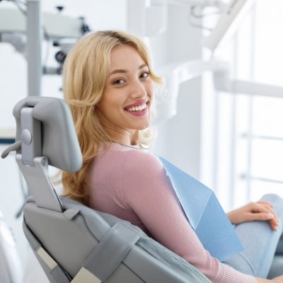 Smiling and relaxed young woman sitting at dental chair in modern clinic, waiting for her dentist. Happy attractive blonde lady attending luxury dental clinic, bacl view, copy space