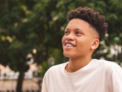 A boy with braces smiling outside in front of some trees.