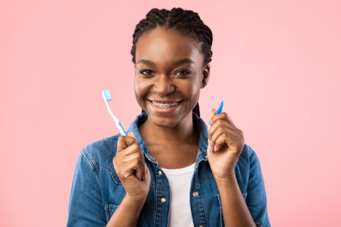 A girl with braces holding up a toothbrush and toothpaste on a pink background.
