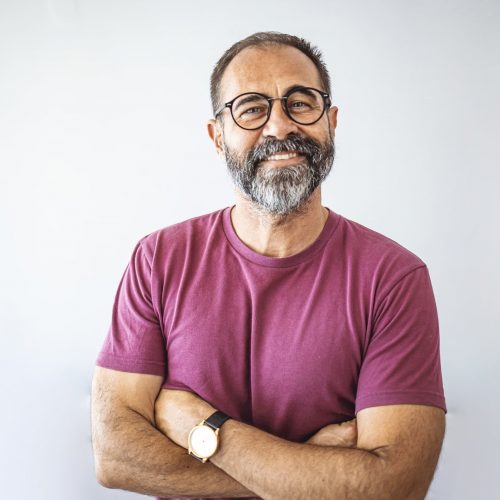 An older man with round glasses standing with his arms crossed in front of a white background