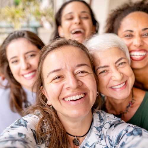 Group of diverse female friends laughing and taking selfies while standing arm in arm together outside