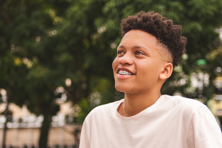 A boy with braces smiling outside in front of some trees.