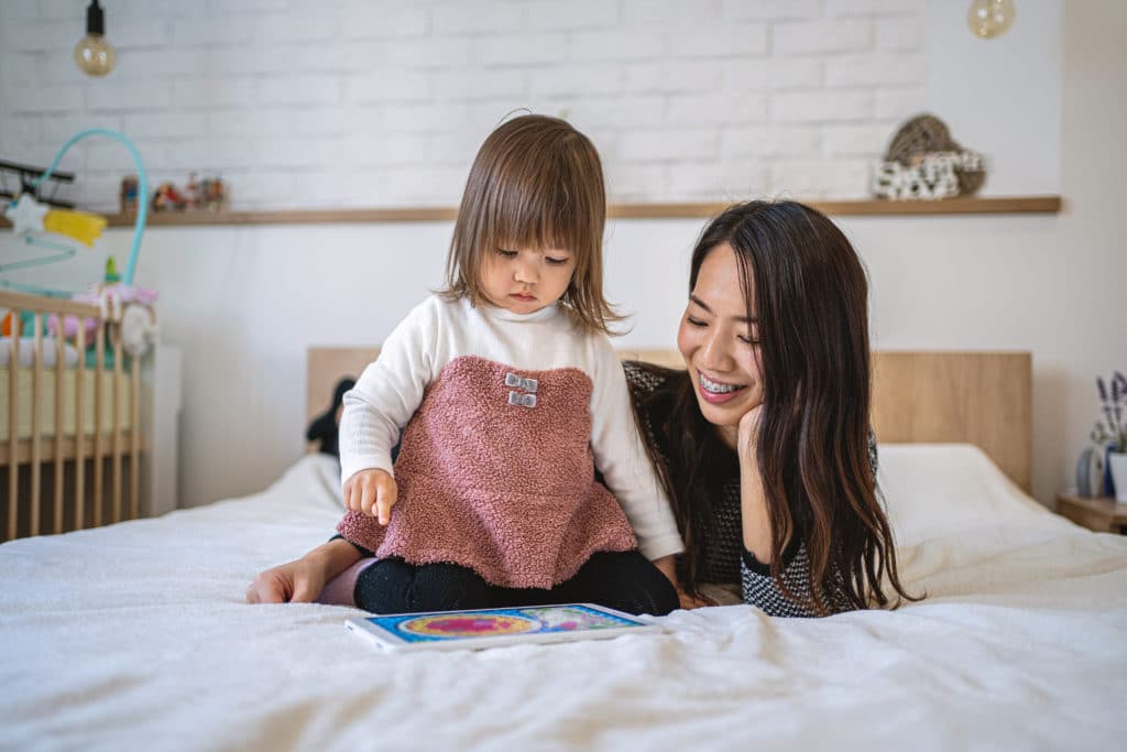 A mom with braces and her toddler lying on a bed and looking at a tablet.