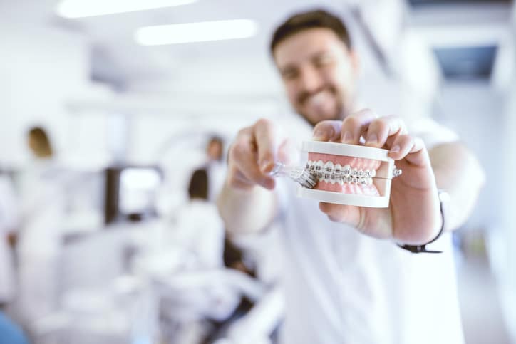 A blurry man holding a model of teeth with braces and demonstrating how to brush them.