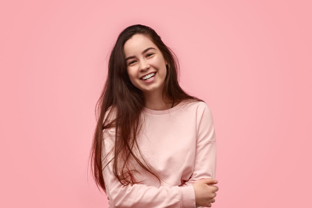 Young woman with long brown hair and an aligned smile in a soft pink sweater with a pink background